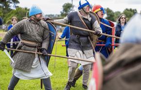 Reenactment of a medieval battle with men in chainmail and helmets holding shields and spears on a grassy field.